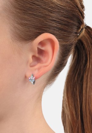 Primo piano di un orecchio con capelli castani raccolti, indossando un piccolo orecchino a cerchio d'argento decorato con un charm a forma di farfalla rosa e blu.