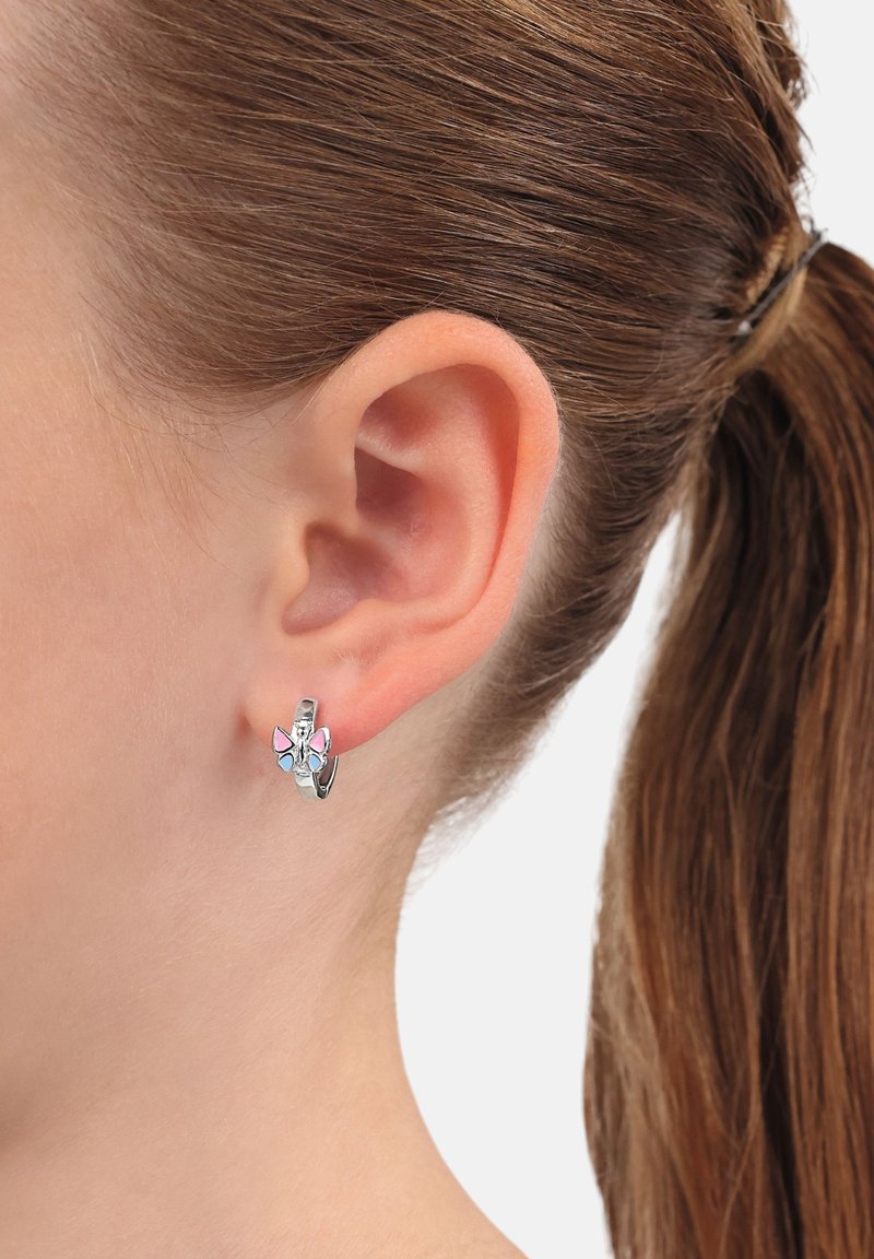 Acercamiento de una oreja con cabello castaño recogido, que lleva un pequeño pendiente de aro plateado adornado con un charm de mariposa rosa y azul.