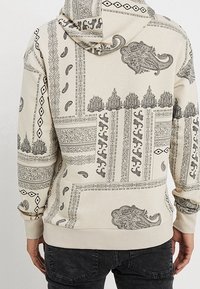 Felpa beige con intricati motivi neri, tra cui design paisley e geometrici. Presenta un cappuccio con cordoncino e polsini a costine.