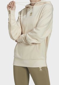 Krämfärgad hoodie med guld bie-logotyp, utrustad med dragkedja på sidan, ribbad fåll och slät textur. Matchas med olivgröna leggings.