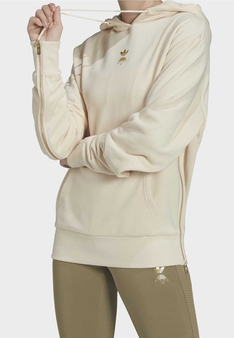 Krämfärgad hoodie med guld bie-logotyp, utrustad med dragkedja på sidan, ribbad fåll och slät textur. Matchas med olivgröna leggings.