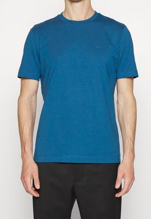Homme portant un t-shirt à col rond bleu uni à manches courtes et un pantalon noir, se tenant les bras détendus le long du corps.