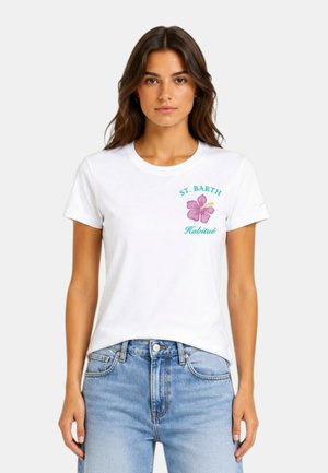 Giovane donna con capelli lunghi e castani indossa una maglietta bianca con un fiore rosa e un testo verde che recita "ST. BARTH Habitué" e jeans azzurri chiari.