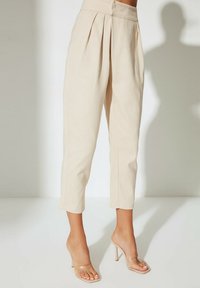 Pantalons beiges ajustés avec plis, longueur courte et texture lisse, associés à des sandales à talons transparents.