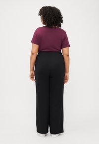 A person with curly hair wears a maroon shirt and black wide-leg pants, facing away against a plain white background.