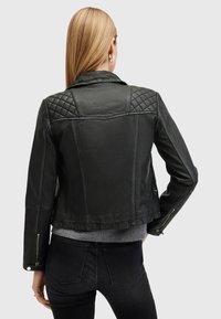 Giacca in pelle nera con dettagli trapuntati sulle spalle, vestibilità slim e polsini con zip, abbinata a un maglione grigio e jeans scuri.