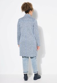 Lång, blå, texturerad kappa med hög krage, sömdetaljer bak och långa ärmar, i kombination med ljusblå jeans och svarta ankelstövlar.