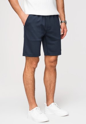 Hombre con pantalones cortos azul marino, zapatillas blancas, camiseta blanca de canalé y reloj de pulsera plateado, de pie con una mano en el bolsillo.