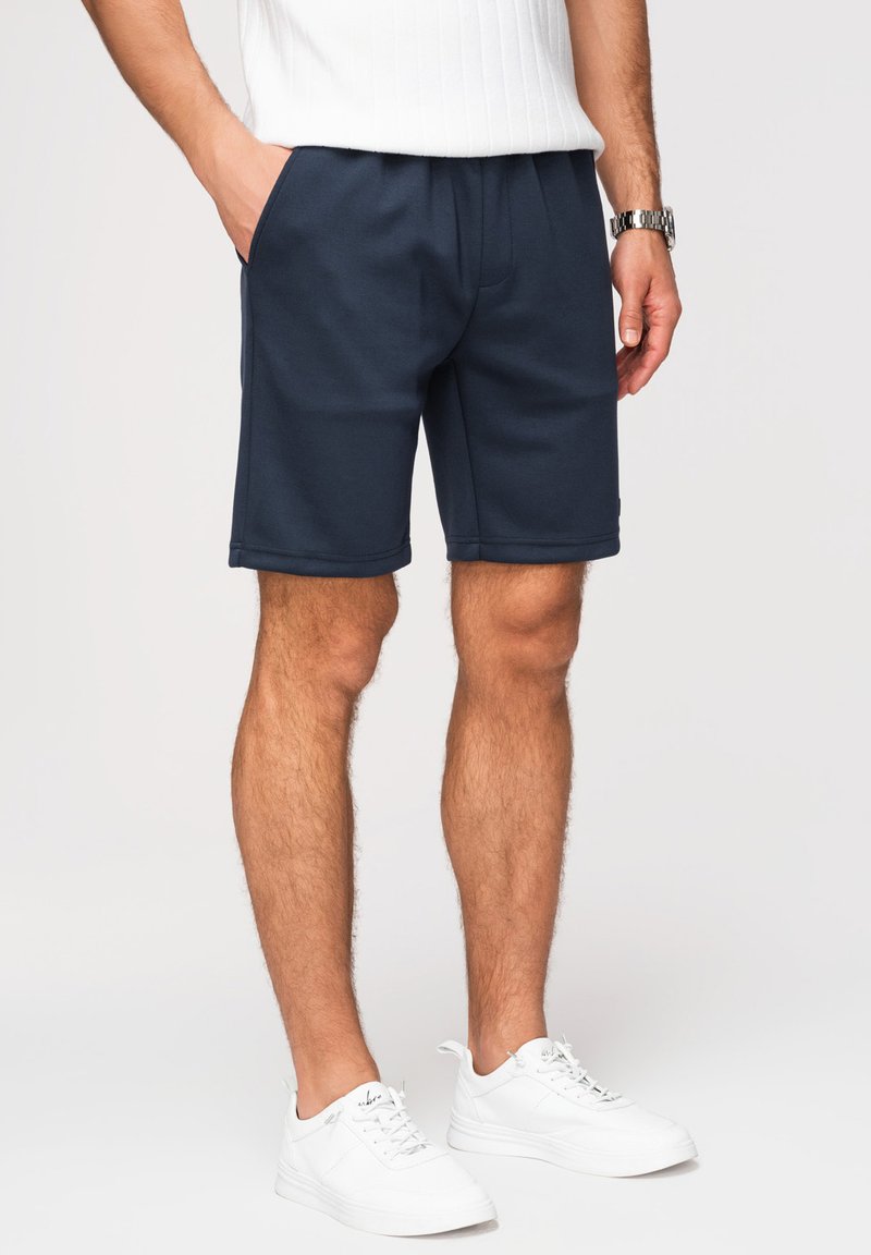 Homme portant un short bleu marine, des baskets blanches, un t-shirt blanc côtelé et une montre argentée au poignet, debout avec une main dans la poche.