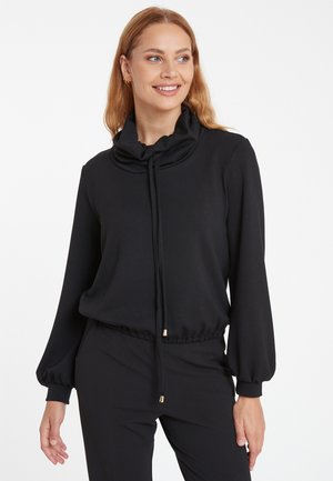 Haut noir à manches longues avec col montant, fermeture à cordon de serrage et ourlet élastiqué. Fabriqué en tissu lisse, il présente des accents dorés.