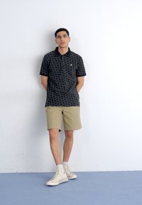 Ung mand står med ryggen mod en hvid væg iført en sort mønstret polo-shirt, beige shorts, hvide sokker og hvide høje sneakers.