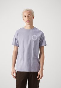 Camiseta de algodón lavanda con manga corta, cuello redondo y detalle gráfico circular en el lado izquierdo del pecho. Modelo vistiendo pantalones marrón oscuro.