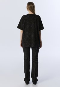 Übergroßes schwarzes Oberteil mit einem strukturierten Leopardenmuster, kombiniert mit schmal geschnittenen schwarzen Hosen mit weißen Nähten und einem ausgestellten Bein.