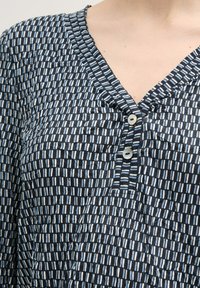 Haut à motifs bleu marine et bleu clair avec un décolleté en V, comportant deux boutons blancs et un tissu texturé avec des rayures horizontales et verticales.