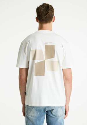 Muž nosiaci biele tričko s béžovými geometrickými tvarmi a malým textom na chrbte, v kombinácii so svetlomodrými džínsami, stojaci chrbtom.