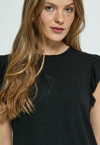 Haut noir à manches courtes avec détails à volants aux épaules, fabriqué en tissu doux. Il dispose d'un col rond et d'une coupe décontractée.