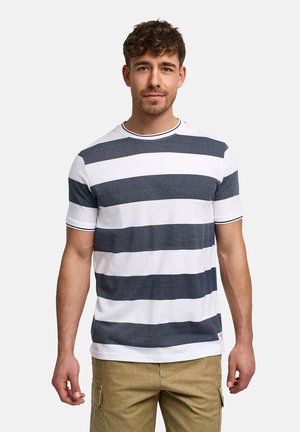 Mann steht mit dem Gesicht zur Kamera, trägt ein weißes und marineblau gestreiftes Kurzarmshirt und beige Cargohosen vor einfachem Hintergrund.