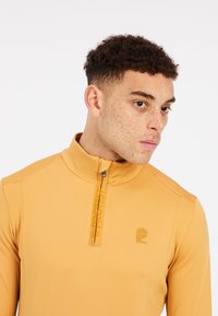 Pulóver s dlhým rukávom v horčicovo žltej farbe, s polozipsovým golierom, hladkou textúrou látky a diskrétnym logom na hrudi.