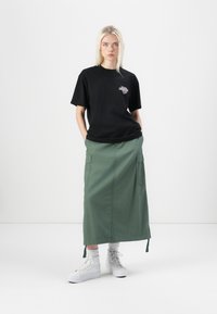 Black cotton t-shirt with a small graphic, paired with a green midi skirt featuring pockets, and white high-top sneakers with textured laces.