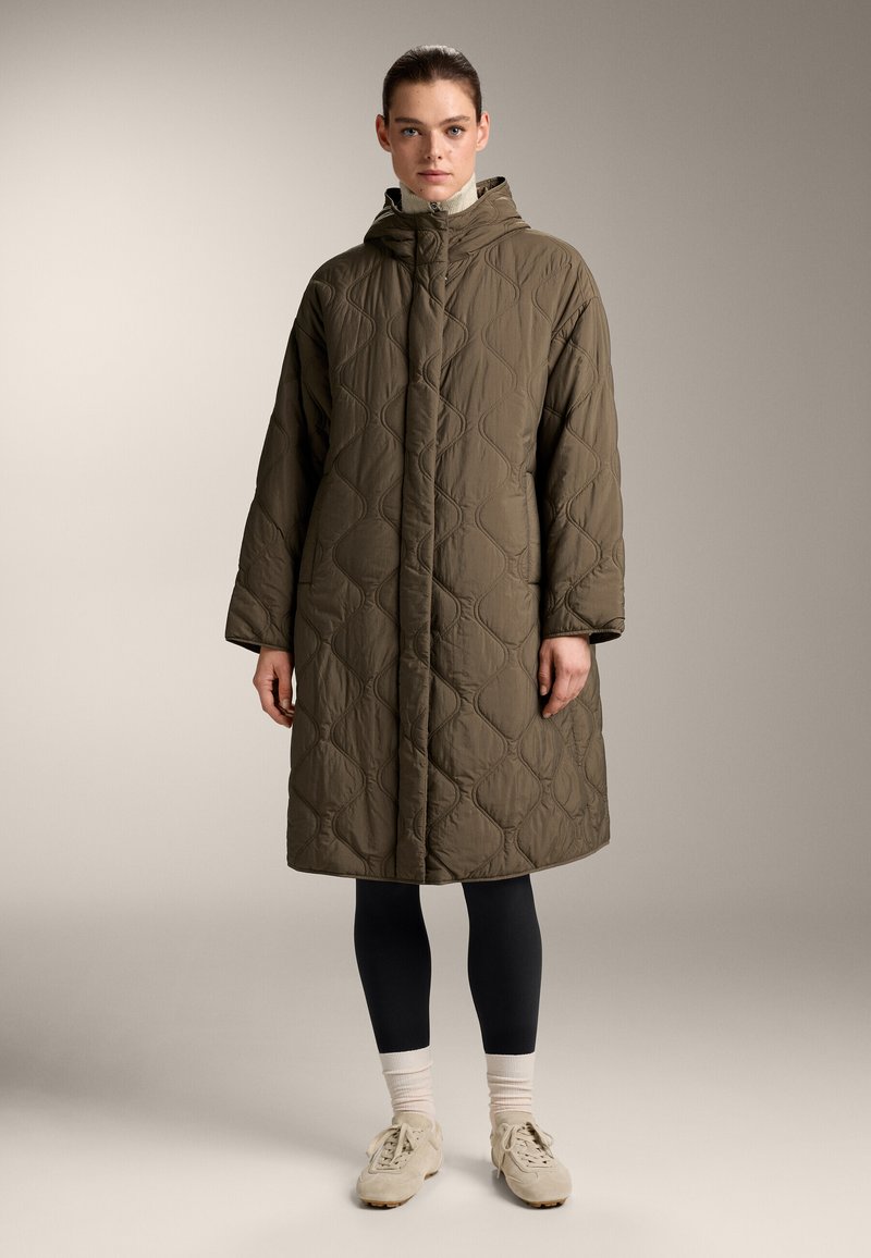 Gevoerde bruine oversized jas met een capuchon, voorzien van een geometrisch patroon, een ritssluiting aan de voorkant en zijzakken, gecombineerd met zwarte leggings en sneakers.