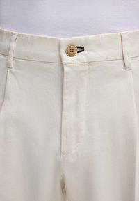 Helle beige Hose aus gewebter Baumwolle, mit Knopfverschluss, Fronttaschen und strukturierter Oberfläche.