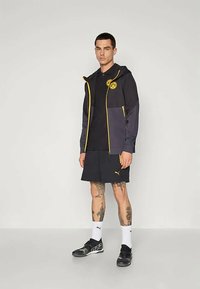 Uomo in piedi che indossa una giacca nera e gialla del BVB, pantaloni corti neri con logo Puma, calze bianche Puma e scarpe sportive nere.