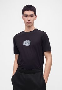 Jeune homme portant un t-shirt noir BOSS et un pantalon noir, les mains dans les poches, debout sur un fond blanc uni.