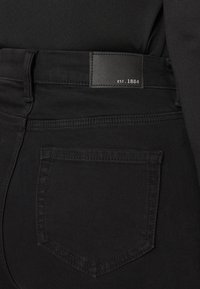 Jeans de mezclilla negra con un parche de cuero rectangular que dice "EST. 1884" y un bolsillo trasero con un diseño y costura estándar.