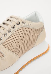 Baskets en suede beige avec des détails en maille perforée, œillets en métal ronds et col rembourré blanc. Présente également une semelle extérieure en caoutchouc orange.
