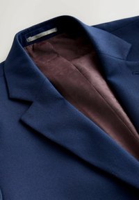 Tengerészkék blézer strukturált szövettel. Belsejében bordó bélés geometriai mintával. Bevágott gallérok és strukturált vállak.