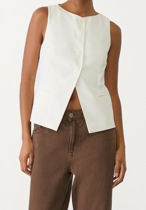 Veste blanche sans manches boutonnée associée à un pantalon large marron taille haute sur une personne debout, les bras détendus.