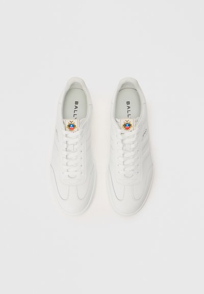 Adidași din piele albă cu un finisaj neted, vârf rotund, șireturi și o patch decorativă cu un emblema colorată pe limbă.
