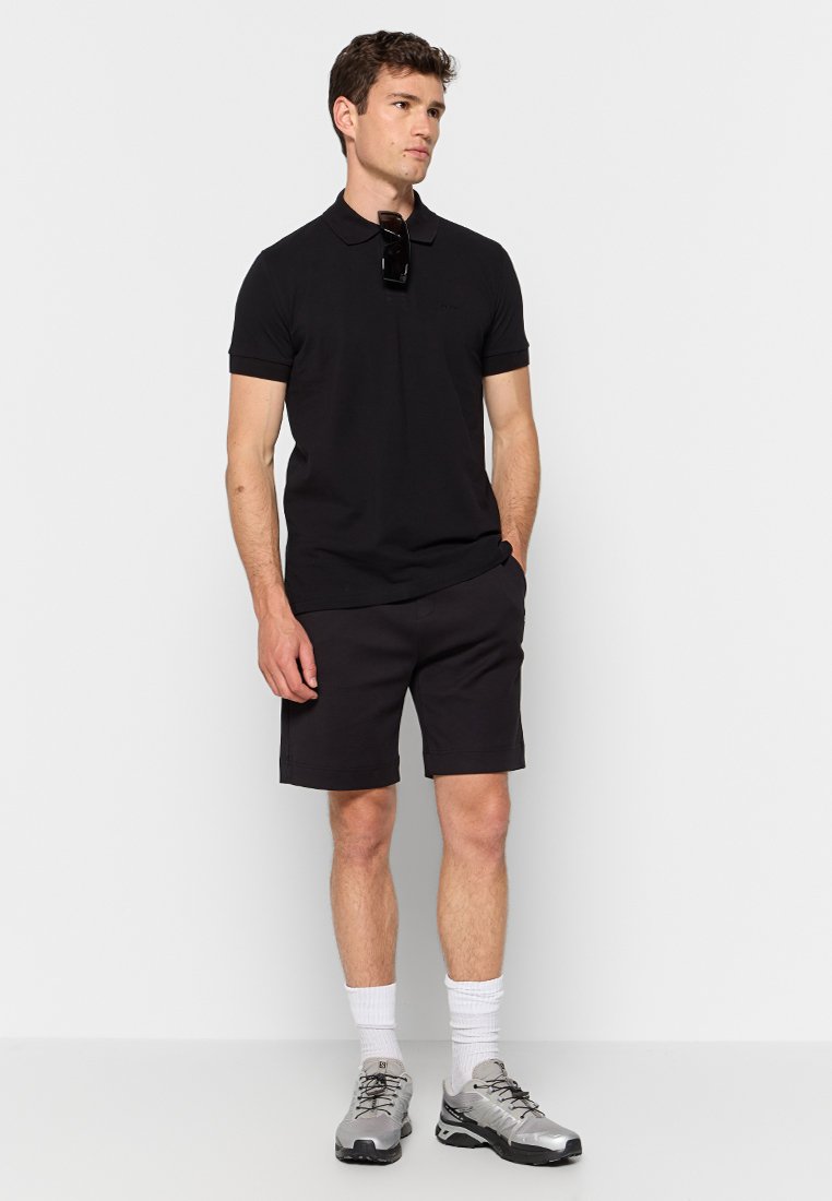 Chemise polo noire, shorts foncés et chaussures de sport grises. Des lunettes de soleil pendent du col. Tissu lisse avec une coupe ajustée.