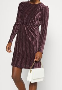 Mujer con un vestido de terciopelo color ciruela, de manga larga y tablas, sosteniendo un bolso blanco con una cadena dorada en un fondo sencillo.