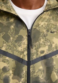 Grøn camouflage zip-up jakke med en tekstureret overflade, sorte accenter og digitale mønstre, lagd over en hvid skjorte.