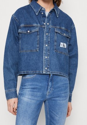 Mujer con chaqueta vaquera corta con botones a presión y bolsillos con solapa, combinada con unos vaqueros de talle alto y color azul desgastado.