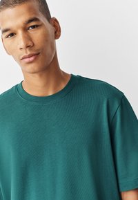 Tamsiai žalsvai mėlynas medvilninis marškinėlis su apvalia iškirpte ir trumpomis rankovėmis, pasižymintis lygiu paviršiumi ir laisvu siluetu. Minimalistinis dizainas be raštų.