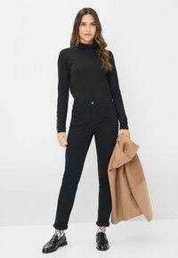 Zwarte langemouwen top, hoge zwarte jeans en zwarte instappers. Beige jas over één arm gedrapeerd. Gemêleerde sokken zichtbaar. Eenvoudige achtergrond.