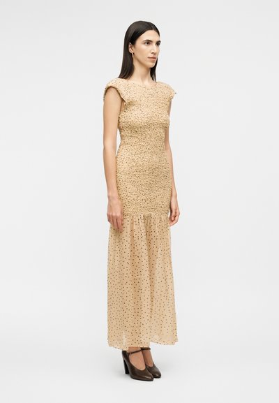 Γυναίκα με ίσια σκουρόχρωμα μαλλιά που φοράει μια μπεζ υφασμάτινη αμάνικη φόρεμα με διαφανές πουά φούστα και μαύρες ψηλοτάκουνες γόβες, στέκεται πλάγια.