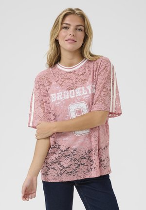 Blonde Frau, die ein pinkes Spitzen-T-Shirt mit der Aufschrift "Brooklyn" und weißen Streifen an den Ärmeln trägt, kombiniert mit dunklen Jeans, vor einem schlichten grauen Hintergrund.