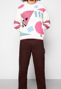 Persona vistiendo un suéter blanco con formas abstractas coloridas y pantalones burdeos, de pie contra un fondo claro y liso.