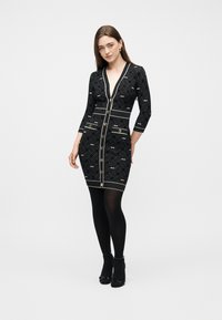 Γυναίκα που φοράει μαύρο εφαρμοστό φόρεμα με λευκά γεωμετρικά σχέδια και χρυσά κουμπιά, συνδυασμένο με μαύρες κάλτσες και μαύρα τακούνια, στέκεται σε λευκό φόντο.