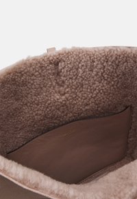 Interno della borsa foderato in shearling marrone con una tasca in pelle liscia. Texture morbida e design minimalista; dettaglio del marchio in oro discreto.