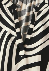 Blusa bianca e nera con audaci motivi a righe, scollo a V e cuciture a contrasto. Il materiale appare liscio e leggero.