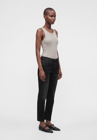 Γυναίκα που στέκεται, φορώντας ένα ανοιχτό γκρι αμάνικο ριμπ μπλουζάκι, μαύρα τζιν ψηλόμεσα και μαύρες μυτερές Mary Jane μπαλαρίνες σε απλό φόντο.