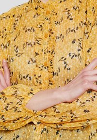 Blusa floreale gialla con scollatura a balze e chiusura con bottone. Presenta motivi floreali neri e maniche lunghe arricciate. Tessuto morbido e testurizzato.