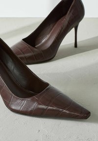 Bruna läderpumps med hög klack och spetsig tå, med ett strukturerat krokodilmönster och en smal klack. Slät inredning.