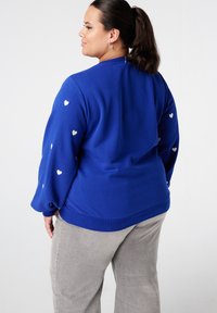 Sweat-shirt à col rond bleu à manches longues, ornée de motifs de cœurs blancs sur les manches. Tissu doux avec une coupe ample et un ourlet côtelé.