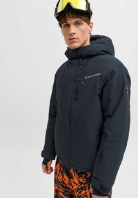 Juoda izoliuota striukė su aukštu apykakle ir reguliuojama kepure, su užtrauktukais ir logotipu ant krūtinės. Dėvima su oranžiniais raštuotais kelnėmis.