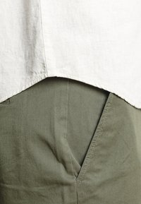 Valkoisen pellavapaidan helma oliivinvihreiden housujen taskun yläpuolella, näkyvissä on kankaan rakenne ja ompelun yksityiskohdat.